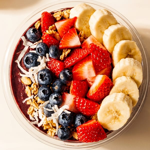 Colorful smoothie bowl with bananas, strawberries, blueberries, granola, and coconut flakes in a clear container.