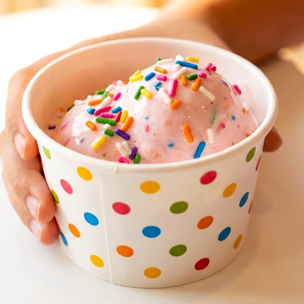 A hand holds a polka-dotted cup of pink ice cream with colorful sprinkles.